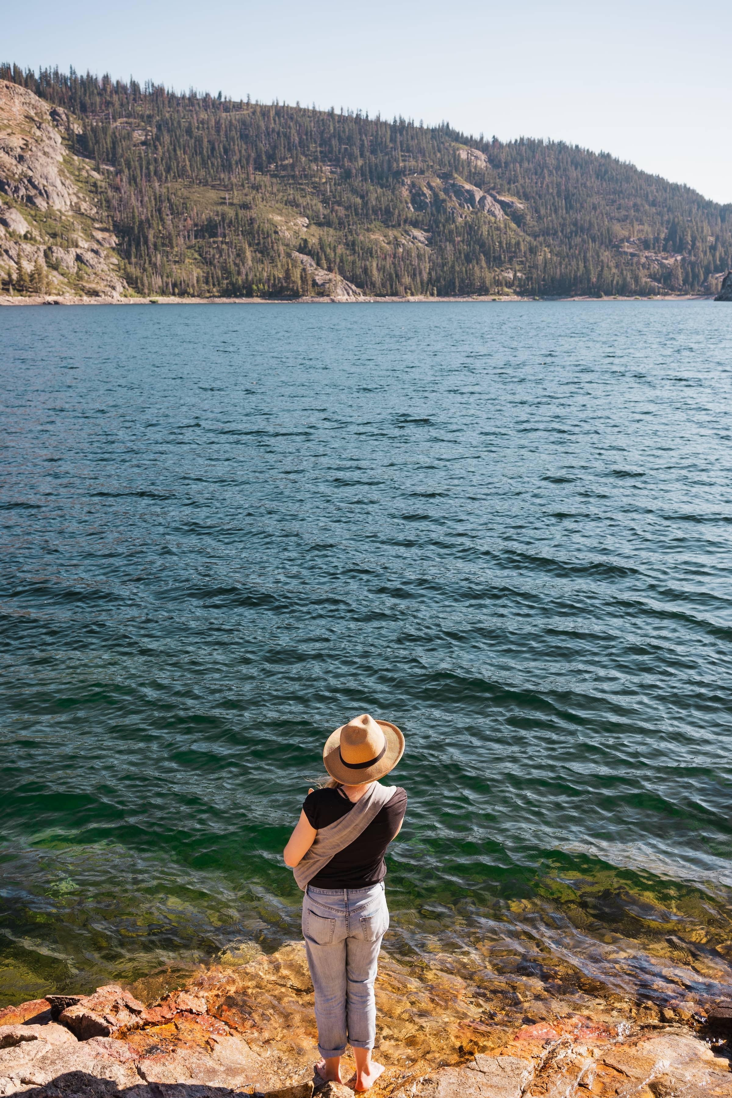 A woman is seen from the back, as she stands at the edge of a lake perched atop rocks, looking out to the distance with mountains on the shore of the other side.