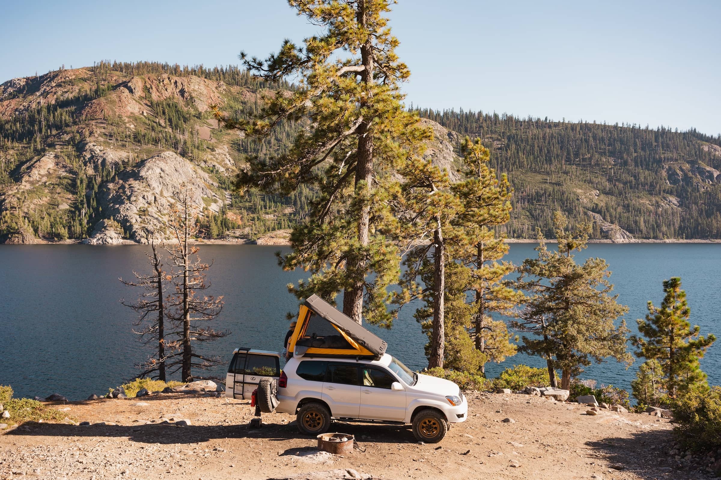 A white SUV with a rooftop tent open on top is seen in profile next to a fire ring, with a lake behind it, and mountains in the distance of the other shore.