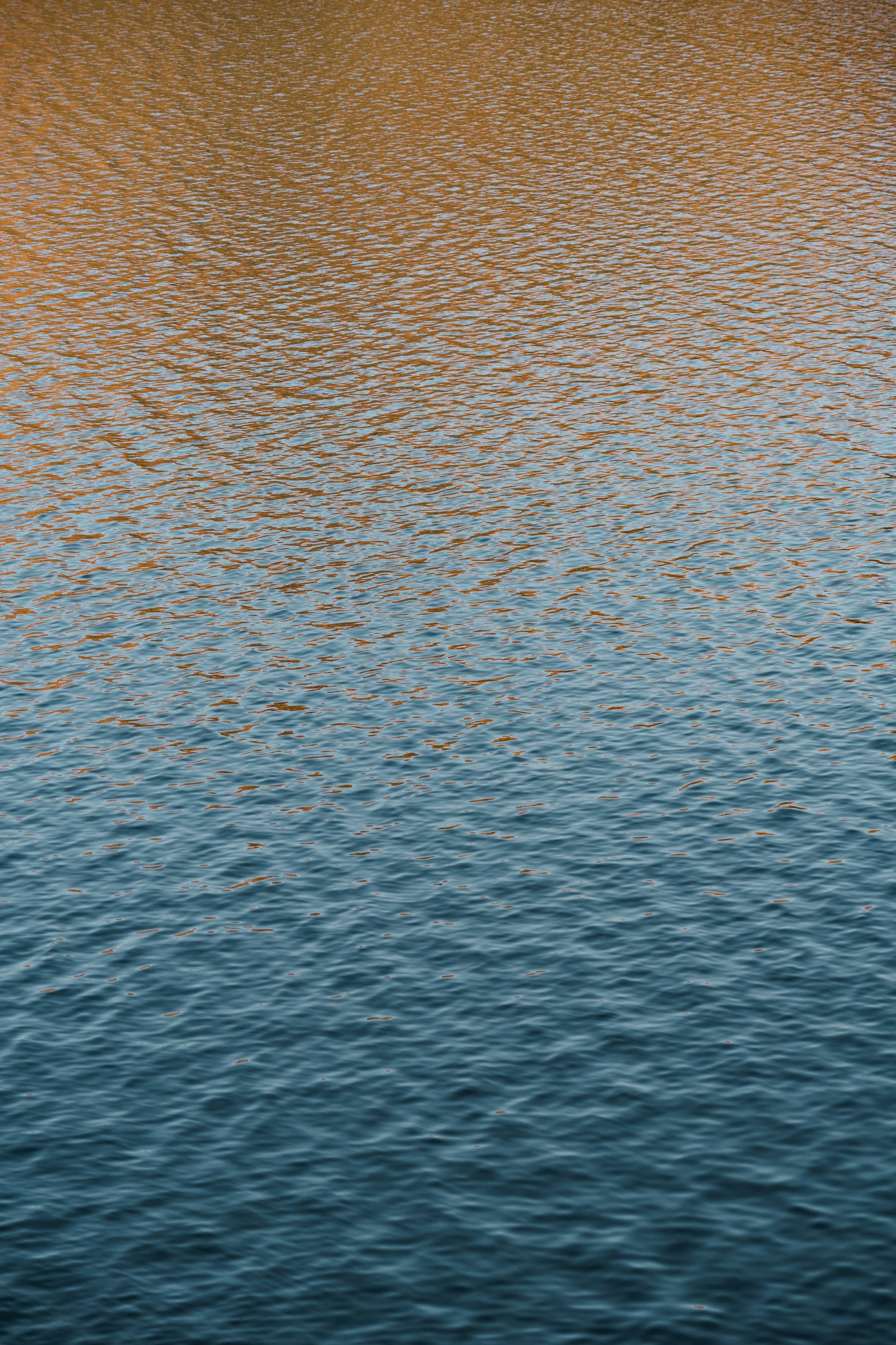 Close up of mildly rippling water with a tint of gold at the top gradienting down to a deep teal at the bottom.