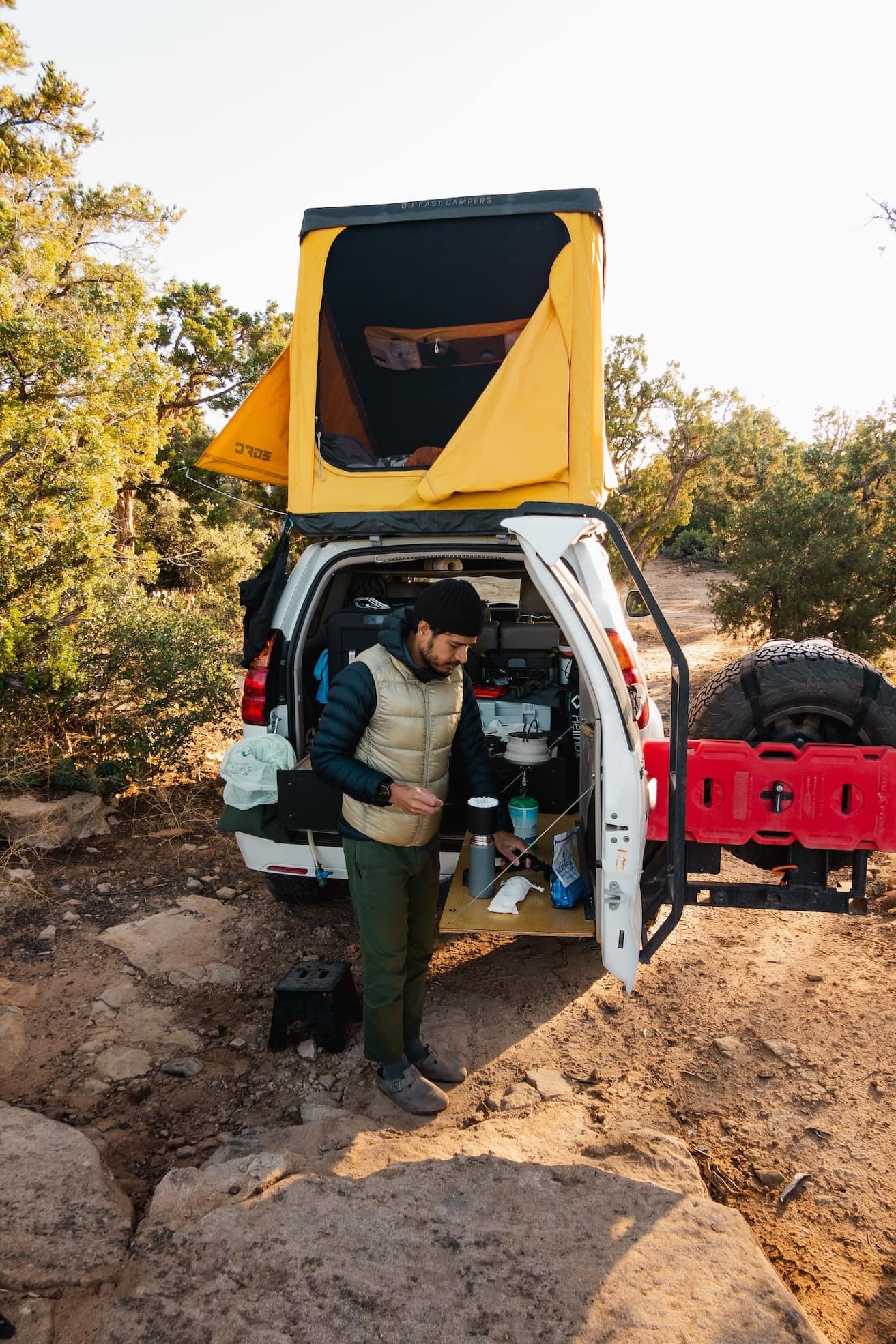 Naz makes coffee out of the back of a white SUV equipped with an open yellow rooftop tent and a rear kitchen setup, in a natural wooded campsite.