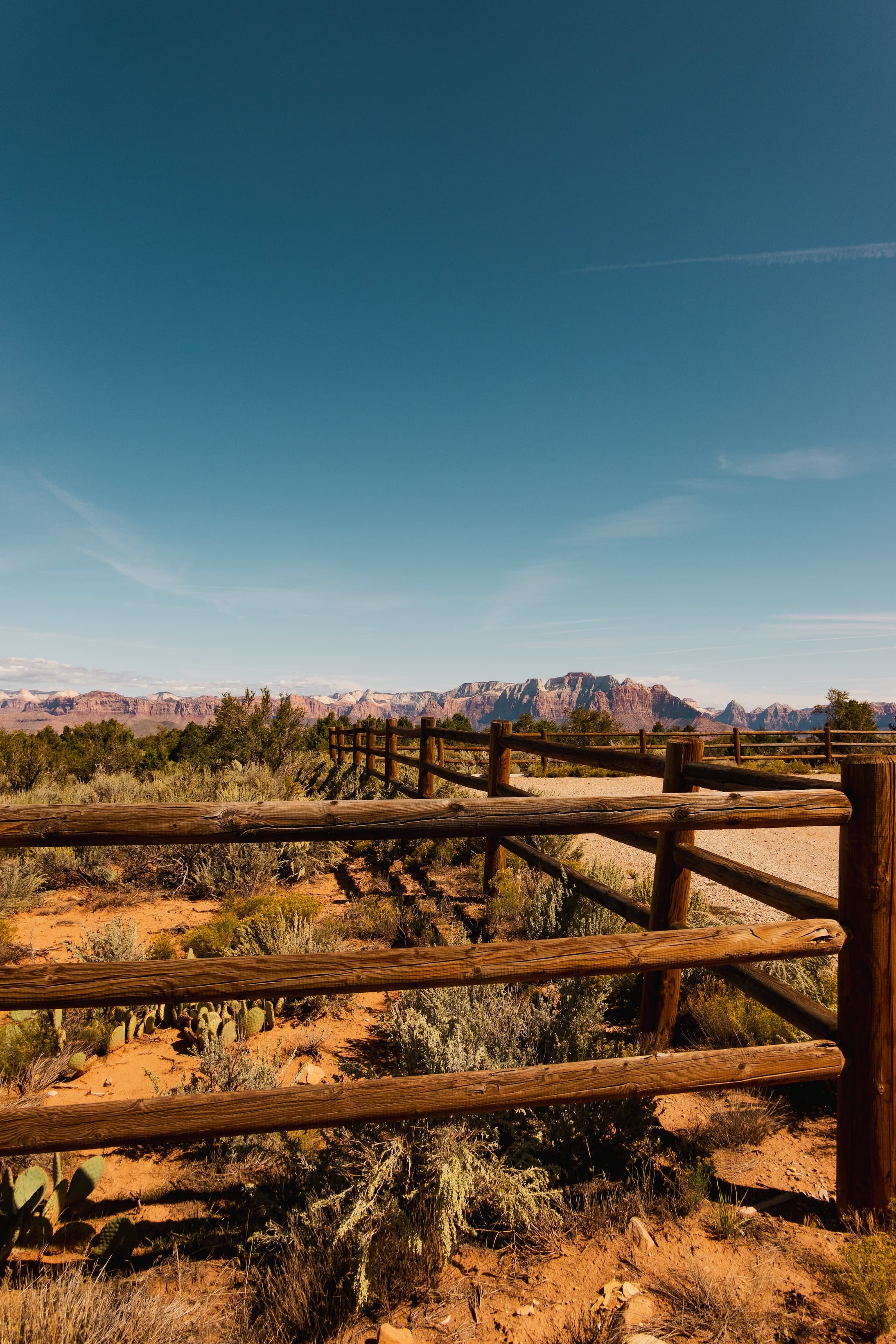 A rustic wooden fence in the foreground frames a panoramic view of distant red rock cliffs under a clear blue sky.