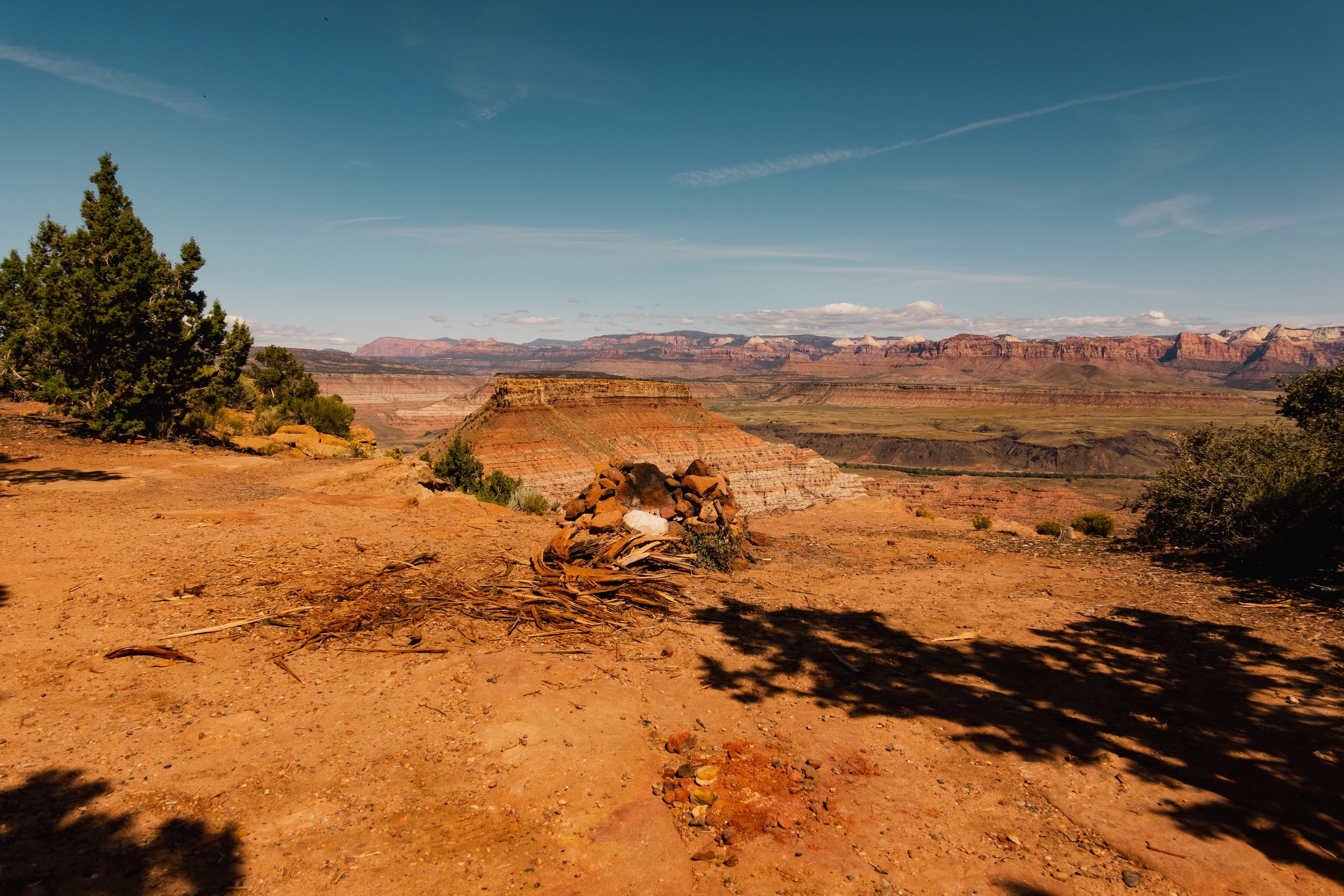 A panoramic view of a vast canyon with layered rock formations in various hues of red and orange, under a wide blue sky with delicate cloud trails. A pile of rocks and wood debris making up a non-active camp fire ring is in the foreground.