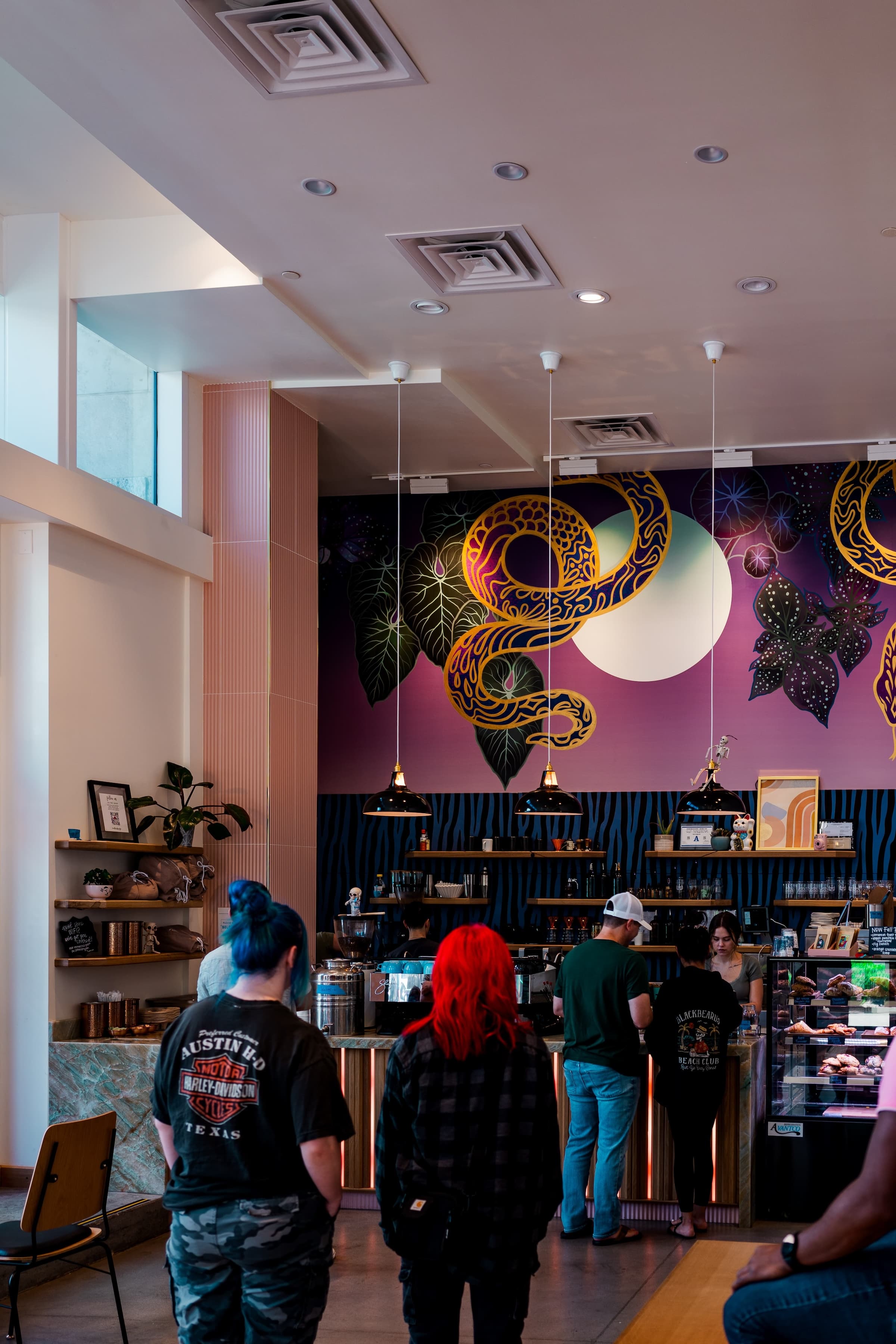 Customers stand in a modern cafe with vibrant cosmic-themed wall art, under a ceiling with recessed lighting and hanging pendant lamps.