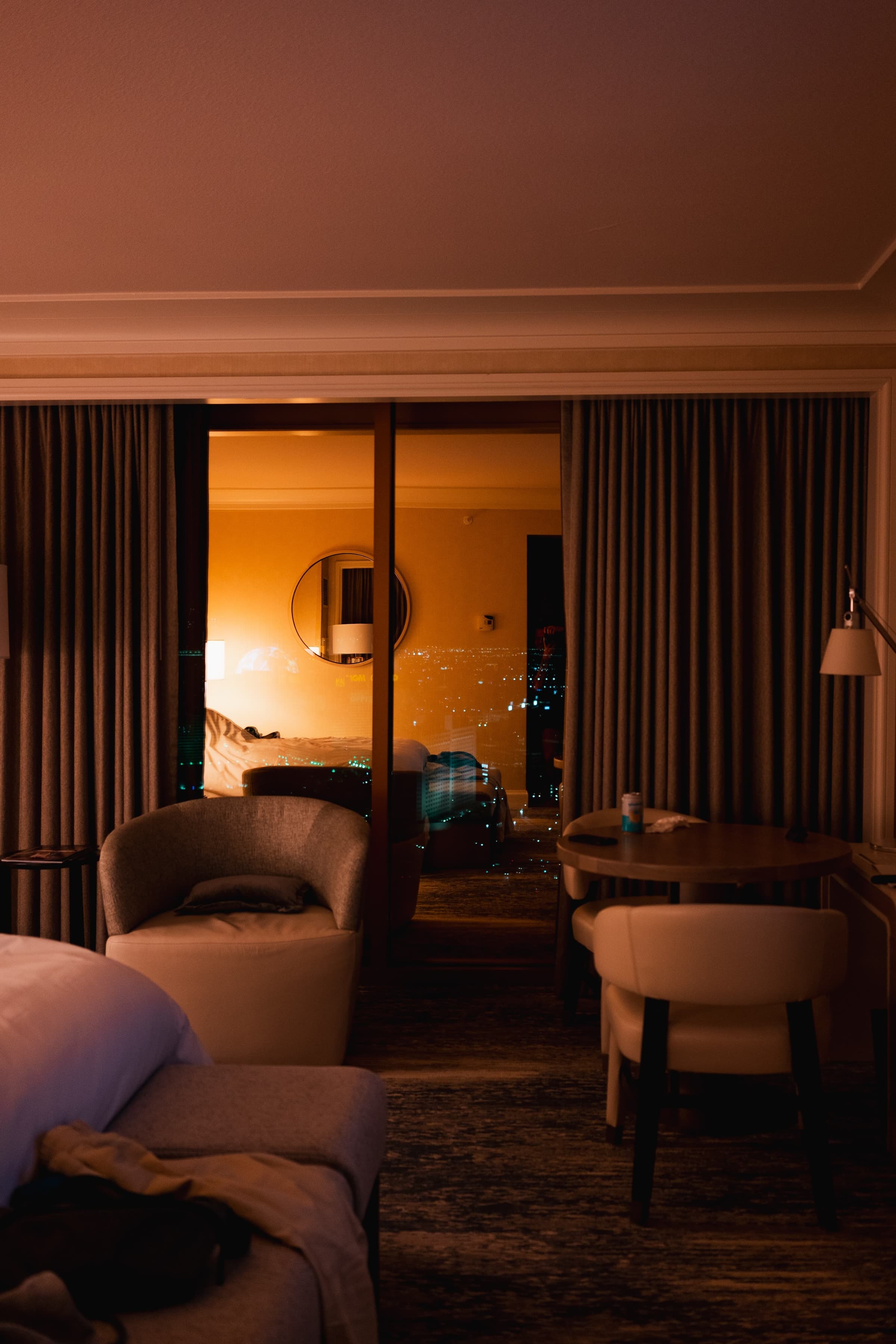 A cozy hotel room interior at night with warm lighting, comfortable furniture, and a large window offering a view of the Las Vegas city lights outside.