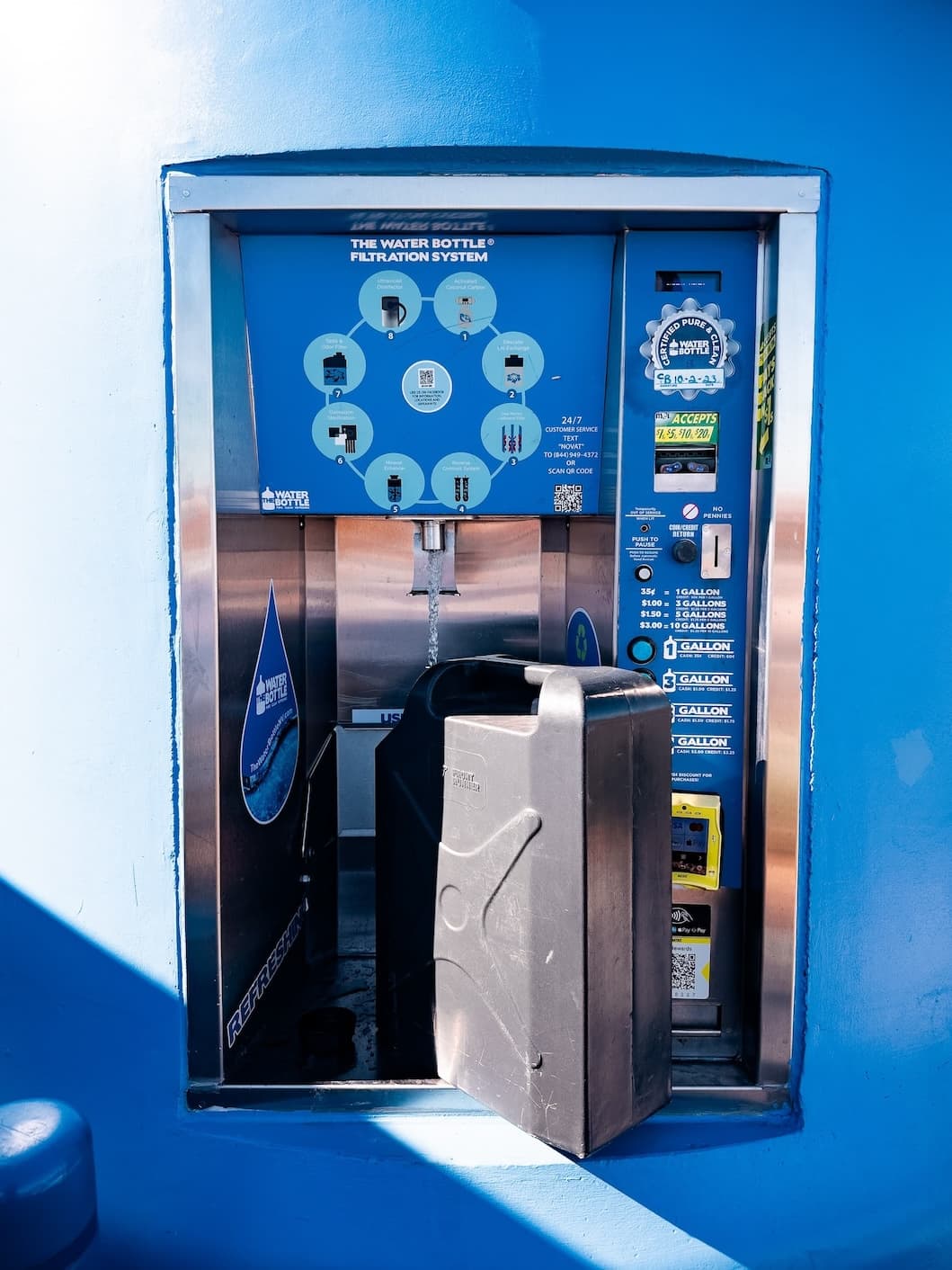 A water bottle refill station with a filtration system set into a bright blue wall, offering various payment options and water amounts.