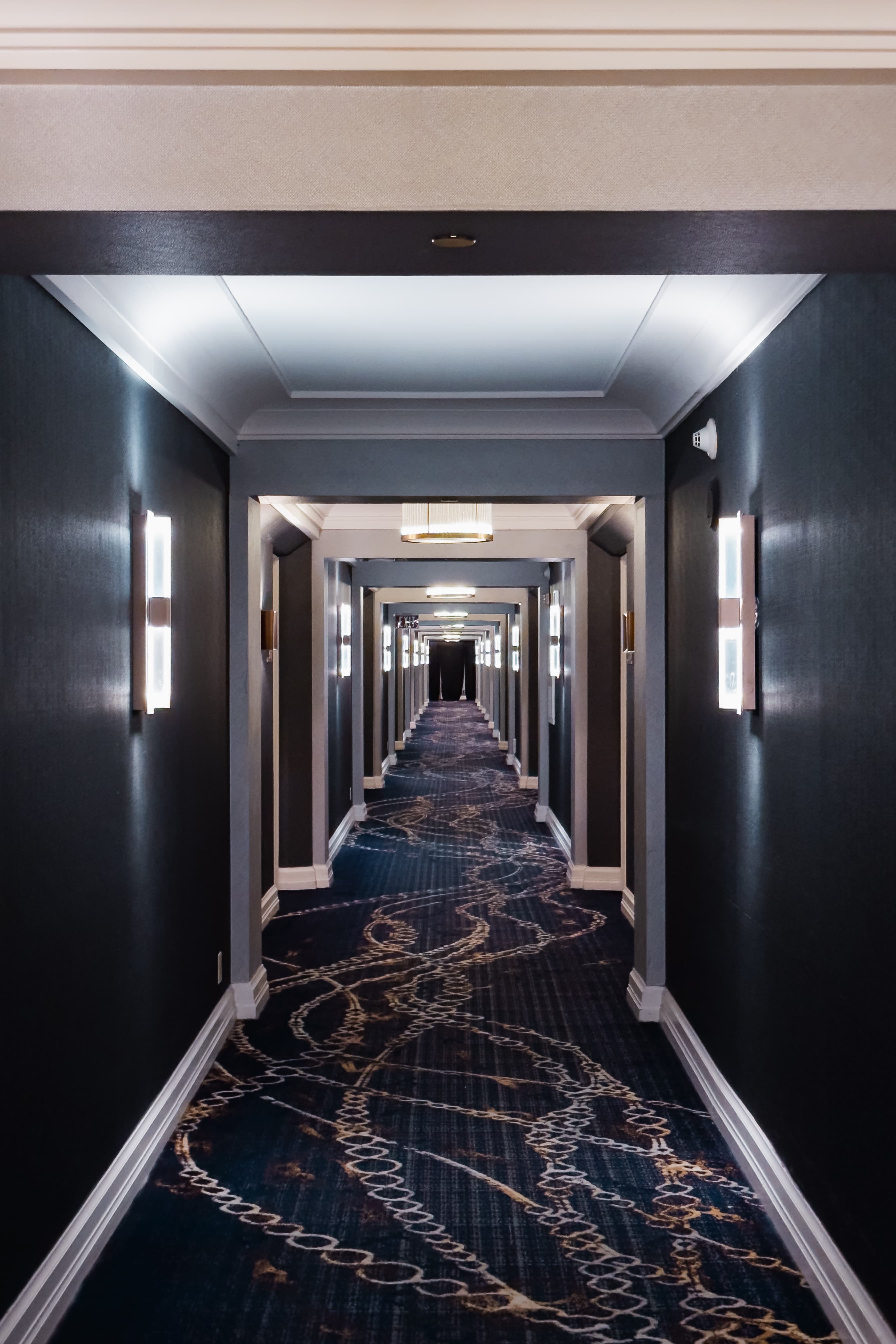 A long, symmetrical hotel corridor with decorative carpet, framed by dark walls and lit by ceiling lights, creating a perspective leading to a vanishing point.