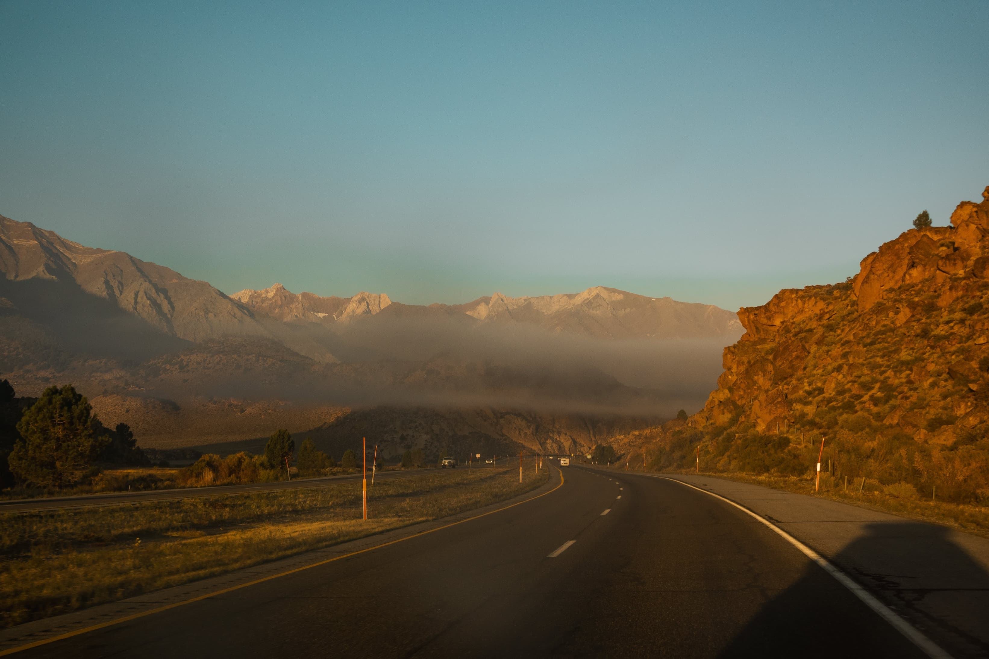 A highway stretches toward mountains veiled in mist, bathed in the golden light of sunrise, with rugged terrain on the right.