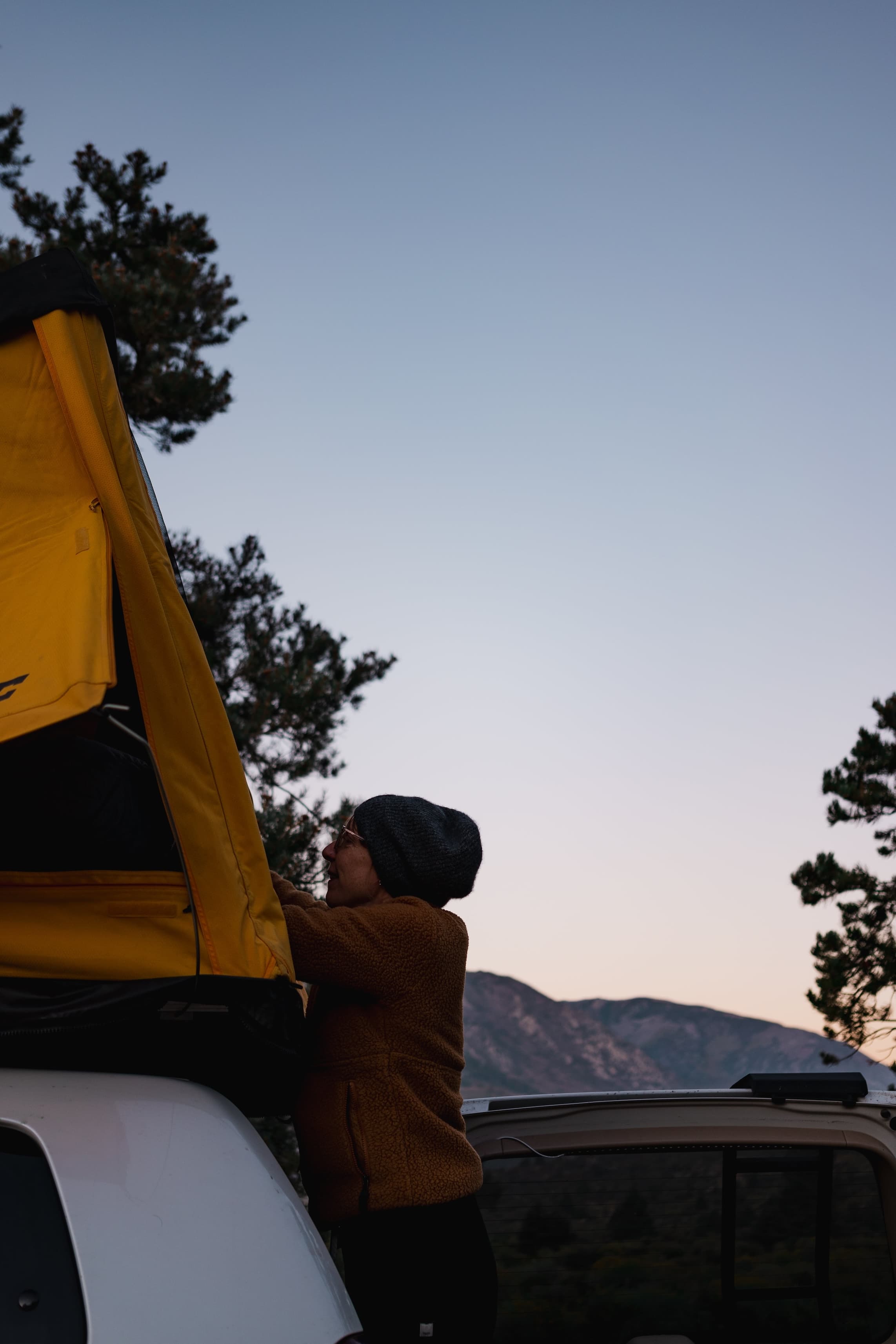 A woman, Jen, in a brown jacket and grey beanie setting up a yellow rooftop tent on a vehicle, with a backdrop of trees and mountains in the twilight.