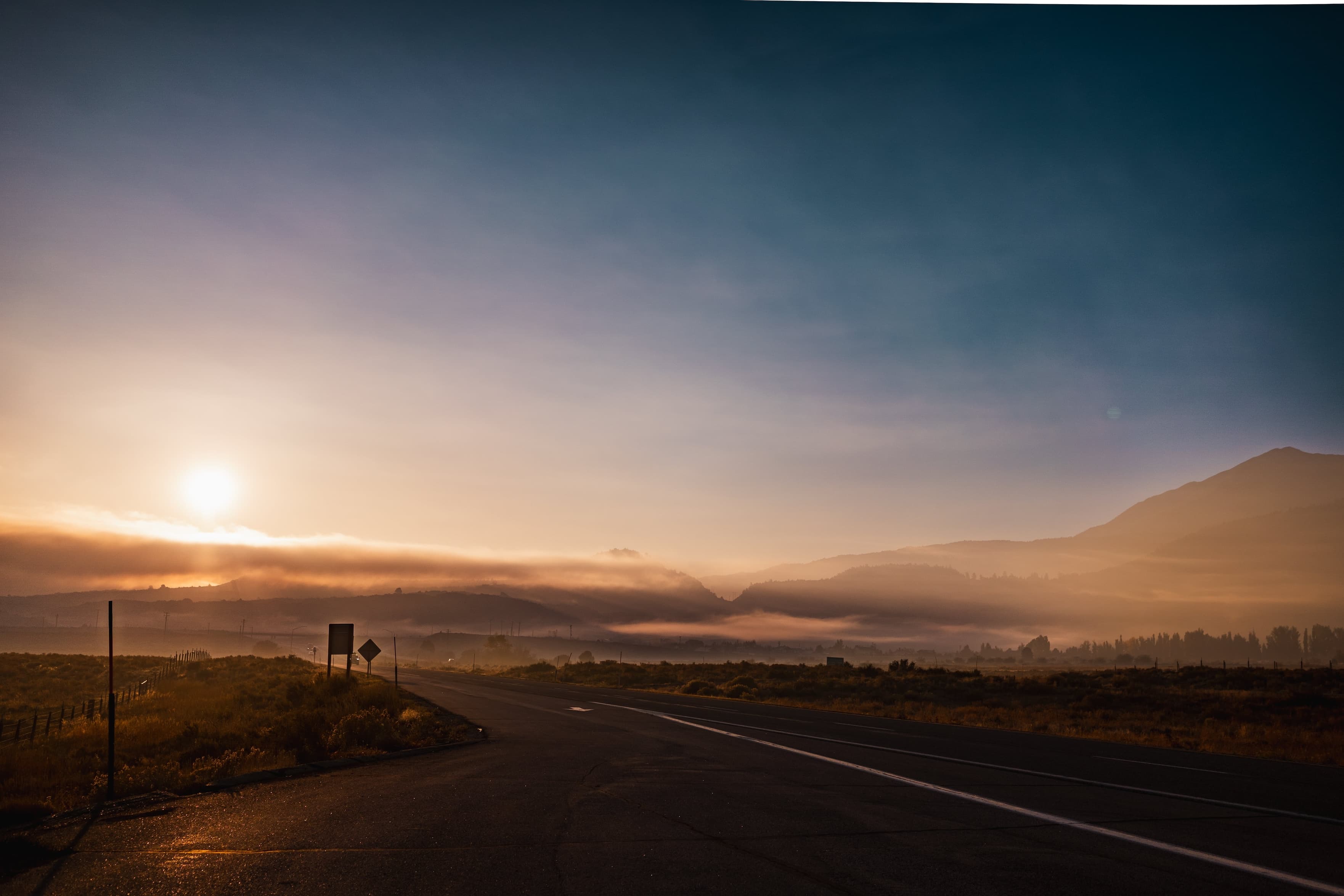A sunrise scene with the sun hanging low over a misty valley, seen from an empty road with open fields on either side.