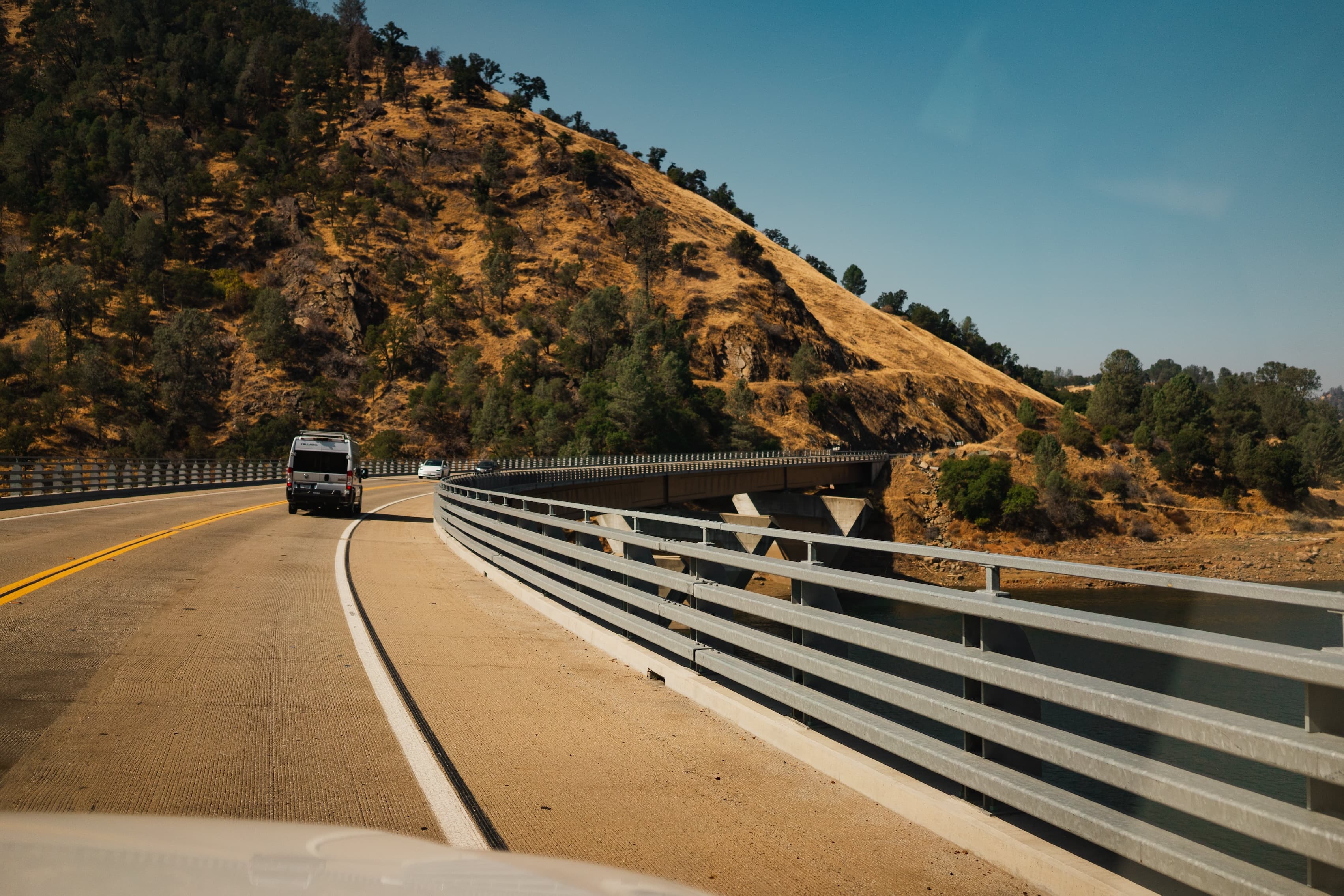 A road curves around a dry hillside with a guardrail on one side and a vehicle driving along, with clear blue skies above.