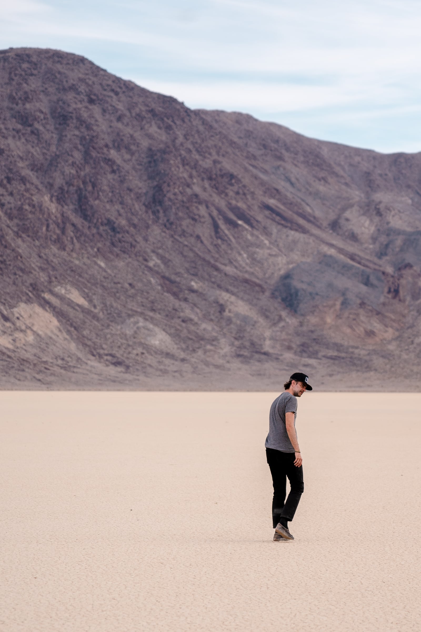 Grandstand Grant, A compressed photo of a tall man in black jeans and black cap with a heather grey tee walking away from the camera but with his head head turned back looking to his right. He's walking on a dry lake bed which is the Grandstand in Death Valley National Park, with a mountain range in the far distance.