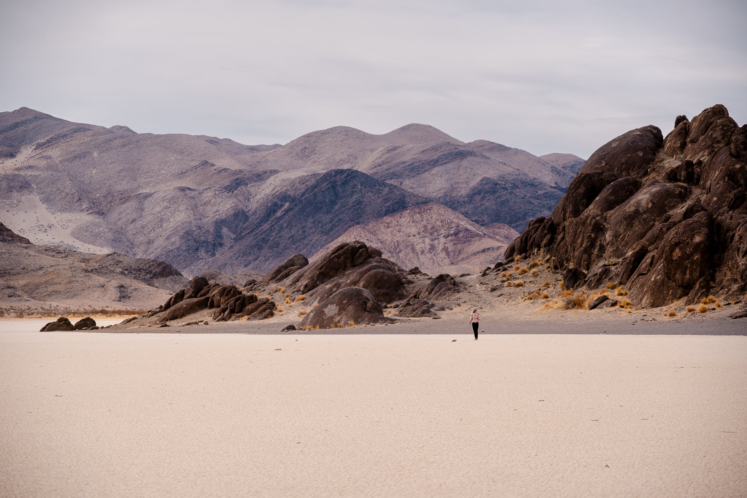 To Comprehend, A woman seen from behind walking in the far distance across a desert playa, dried up with the Death Valley Grandstand formation of rocks in the mid distance, looming in a dark brown, while the Panamint Range of mountains is in the background. 