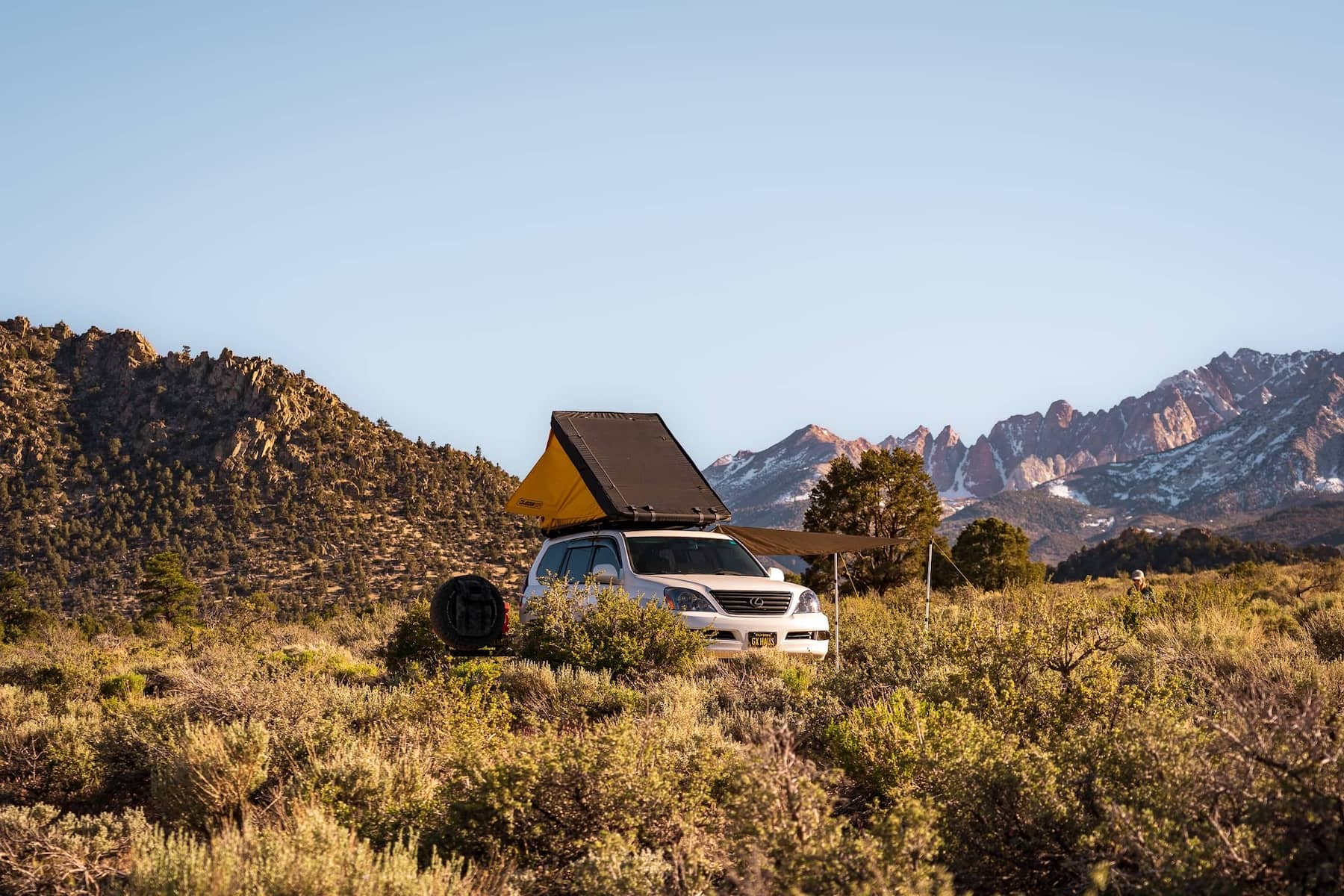 A white SUV with a rooftop tent and an awning extended sits in the middle of some low bush and desert grassland while mountains are seen in the background.