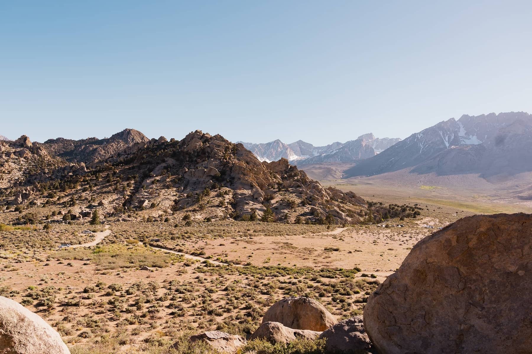 A mountainous desert scene with dirt roads criss-crossing with mountains in the background and large boulder piles where rock climbers are exerting their sport. A few cars are scattered about.