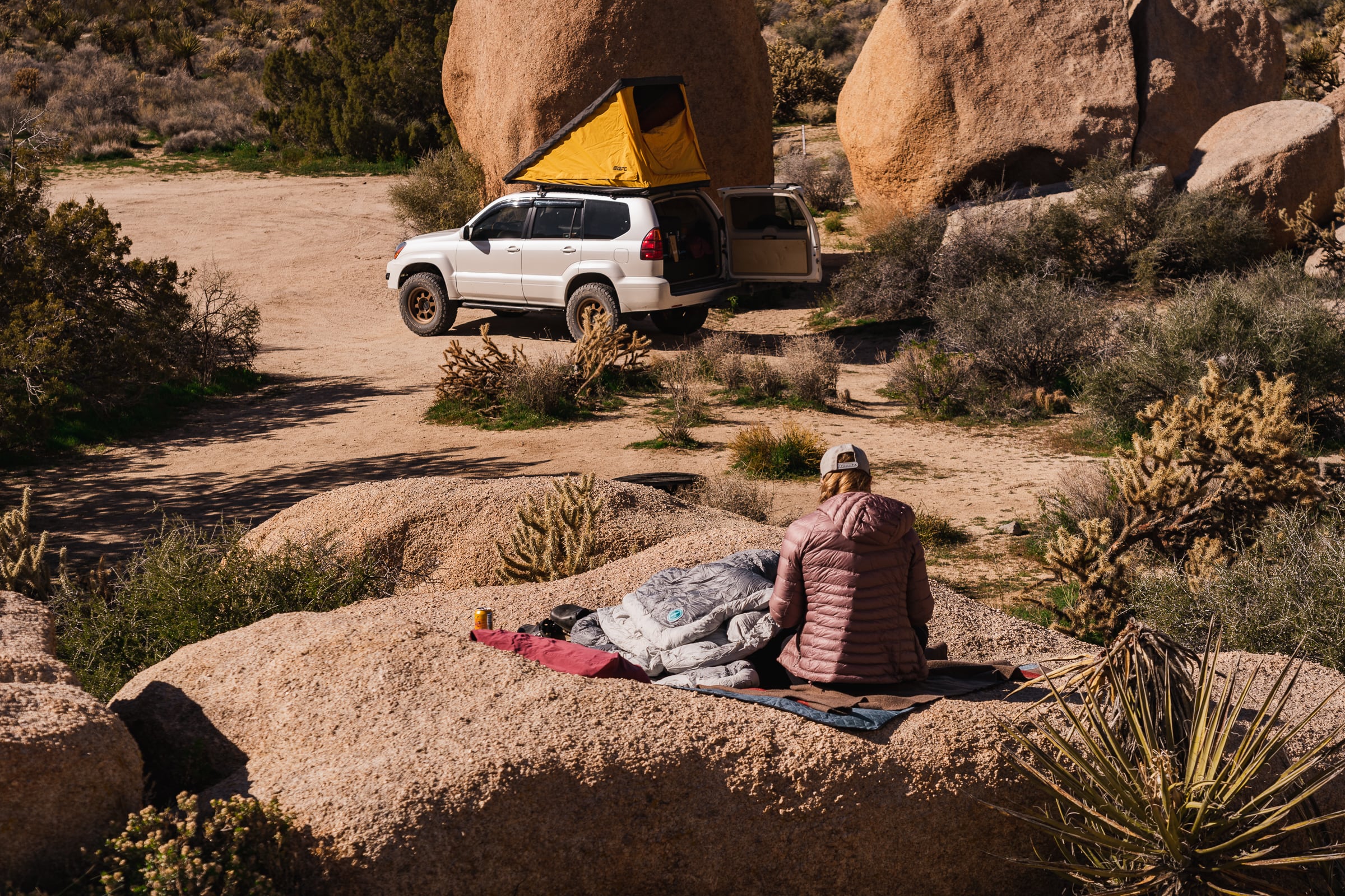 A woman is knitting on a huge rock, set amongst a boulder field, with cactus and other flora surrounding the frame. A 4x4 Lexus GX470 sits off in the distance with a GoFastCampers Superlite rooftop tent open above it, next to its own impressively tall boulder.