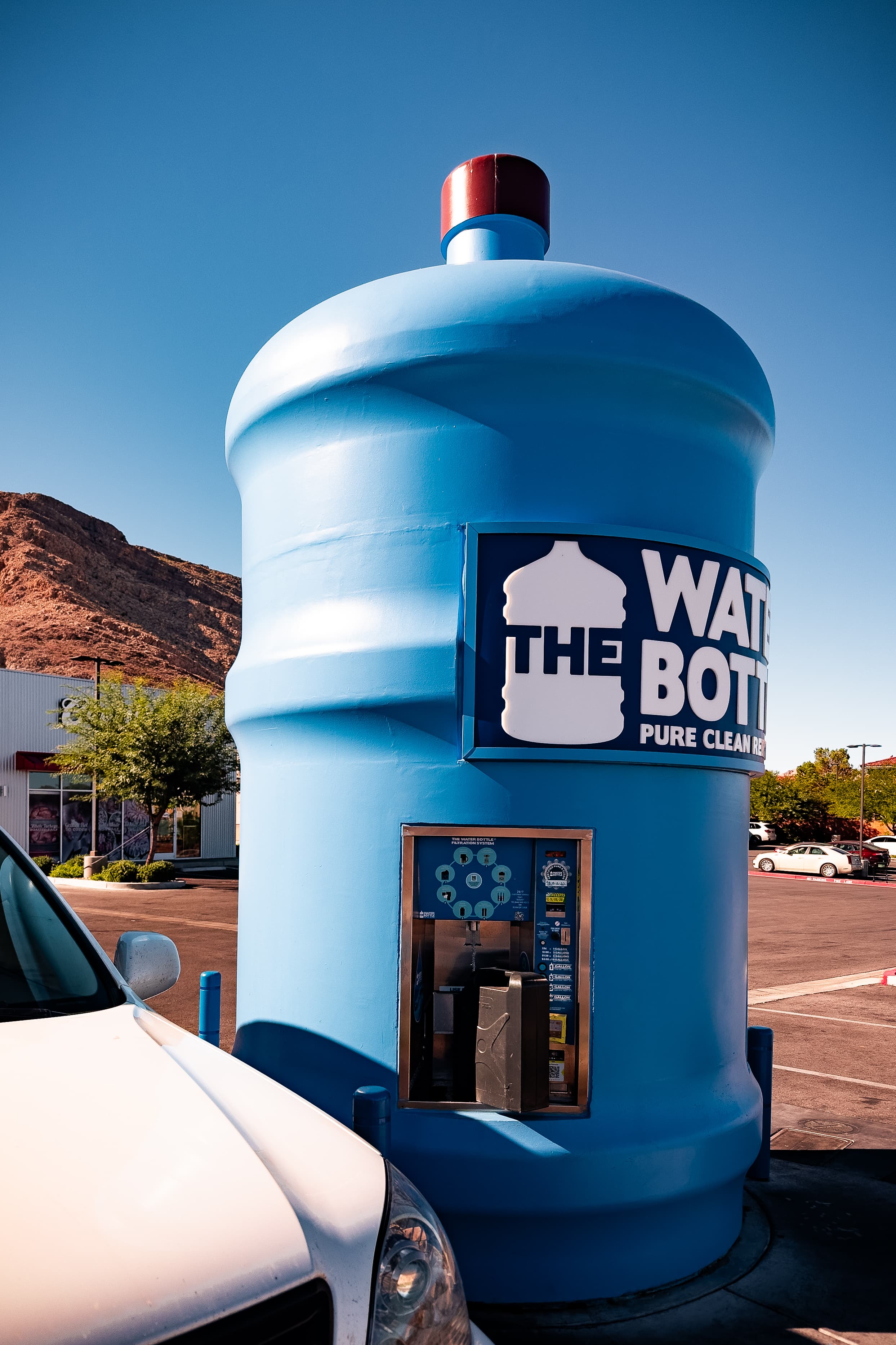 A large blue water bottle-shaped structure housing a water refill station in a parking lot, with clear skies and a mountain in the background.